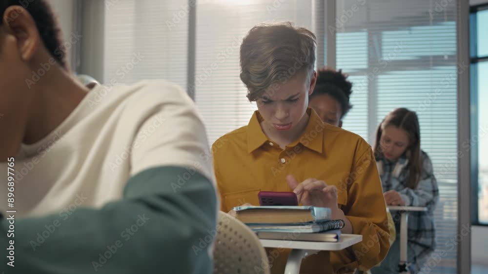 Close-up of cheerful Caucasian teenage boy sitting at desk among diverse classmates at school, cheating at test by using Internet on smartphone during lesson Close-up of cheerful Caucasian teenage boy sitting at desk among diverse classmates at school, cheating at test by using Internet on smartphone during lesson