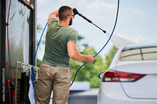 Washing luxury white auto with water gun on an open air car wash. Back view of confident man cleaning his car with high pressure water jet. Technical service concept.