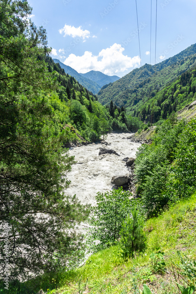 A muddy mountain river flows among trees, rocks and bushes. Huge stones in the riverbed. High voltage wires are visible A muddy mountain river flows among trees, rocks and bushes. Huge stones in the riverbed. High voltage wires are visible