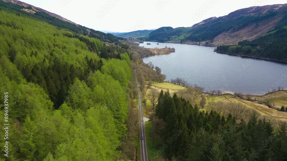 Aerial view of car driving along scenic road through green pine tree forest on Loch Chon in the Scottish highlands, Scotland. Scottish landscape, coniferous forest and rolling mountains above loch Aerial view of car driving along scenic road through green pine tree forest on Loch Chon in the Scottish highlands, Scotland. Scottish landscape, coniferous forest and rolling mountains above loch
