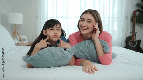 Happy asian daughter and smart mom lie on bed while looking at camera. Energetic family spend time and prepare to sleep together. Cute girl and mom smile to camera show love and affection. Pedagogy.