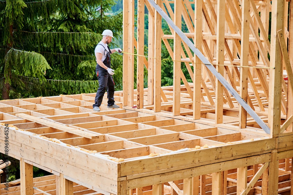 Carpenter constructing house with wooden frame. Man measures distance using tape measure, wearing protective clothing and helmet. Concept of environmentally sound, modern construction. Carpenter constructing house with wooden frame. Man measures distance using tape measure, wearing protective clothing and helmet. Concept of environmentally sound, modern construction.