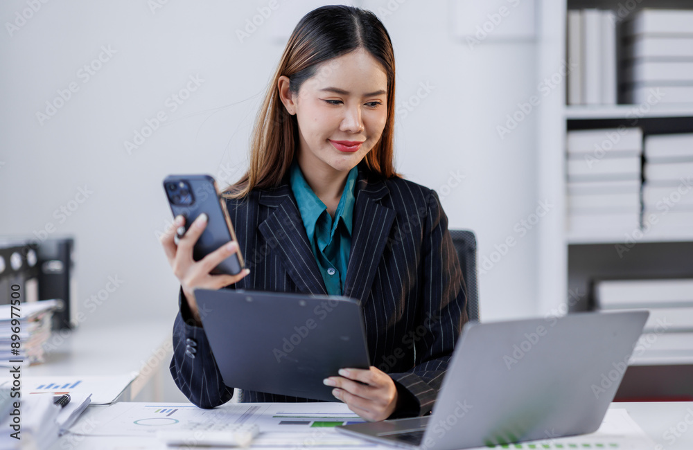 Asian woman sit at workplace desk holds cellphone staring at laptop, synchronize data between computer and gadget in office, use corporate devices and business application, plan work, use organizer Asian woman sit at workplace desk holds cellphone staring at laptop, synchronize data between computer and gadget in office, use corporate devices and business application, plan work, use organizer