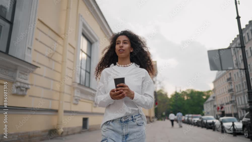 Positive black woman strolling alone in city center in summer day, medium shot. Happy multiracial young lady drinking hot coffee to go and admiring architecture, Arab female tourist in downtown