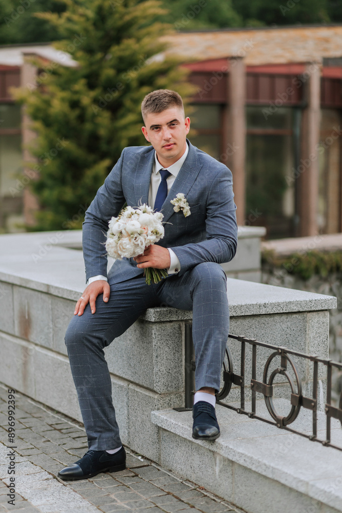 Obraz premium A man in a suit and tie sits on a stone wall holding a bouquet of flowers. Concept of elegance and sophistication, as the man is dressed in formal attire