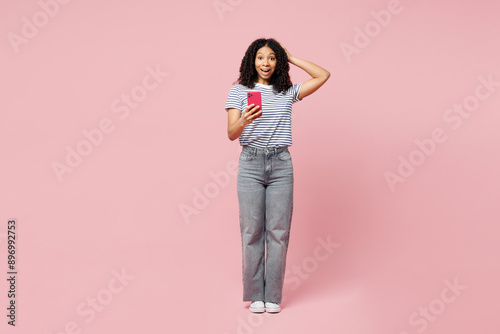 Full body happy surprised shocked fun little kid teen girl wears blue striped t-shirt casual clothes hold head use mobile cell phone isolated on plain pastel light pink background. Childhood concept. © ViDi Studio