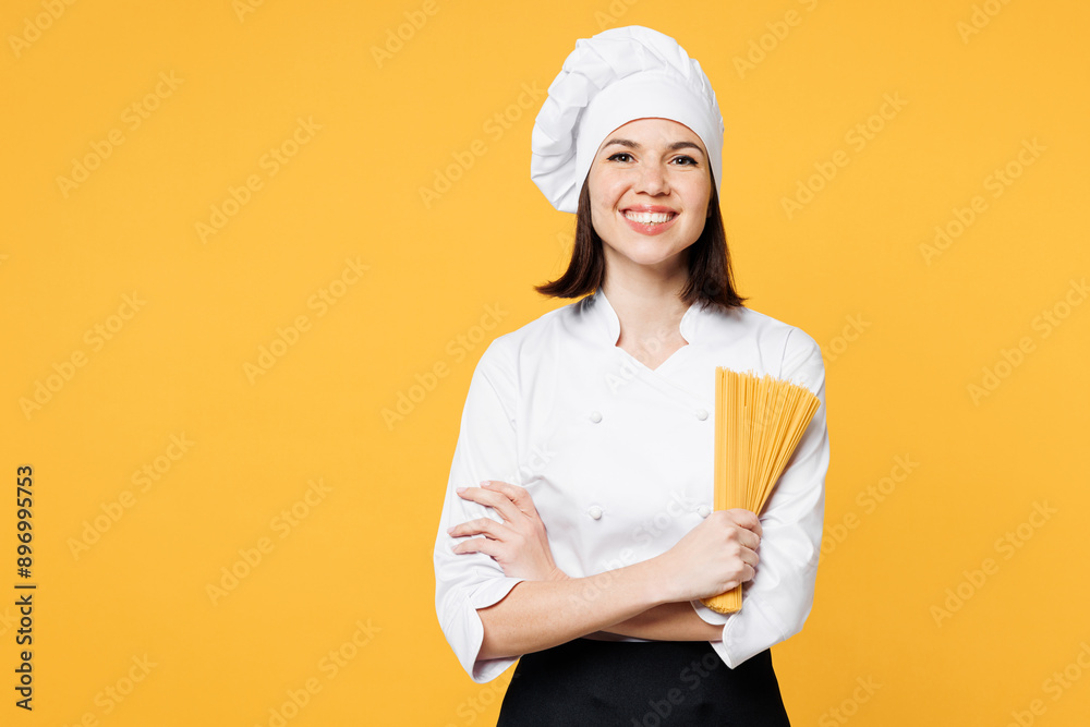 Young chef cook baker woman wears white shirt black apron uniform toque chefs hat holding in hand stack of raw spaghetti look camera isolated on plain yellow background studio. Cooking food concept. Young chef cook baker woman wears white shirt black apron uniform toque chefs hat holding in hand stack of raw spaghetti look camera isolated on plain yellow background studio. Cooking food concept.