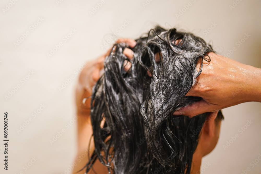A black hair woman is washing hair with shampoo and conditioner during taking rain shower. Healthcare activity scene, Close-up from the back.
