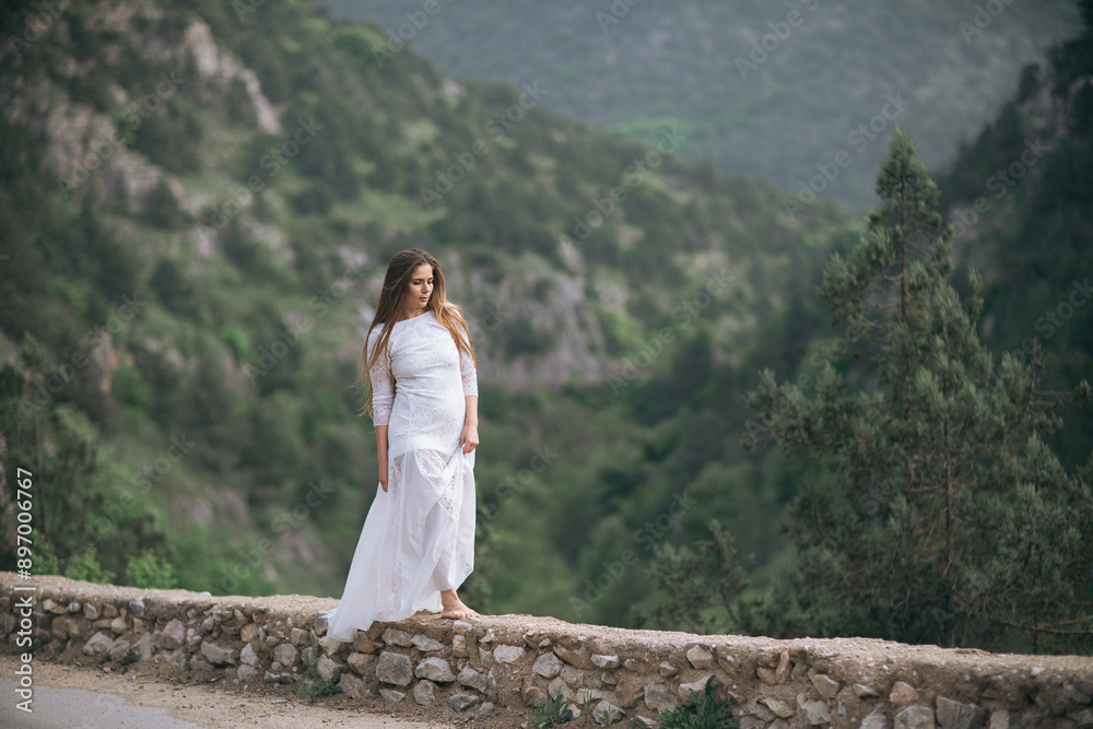 Naklejka premium A woman in a white dress is standing on a stone wall overlooking a mountain. The scene is serene and peaceful, with the woman's dress and the surrounding landscape creating a sense of calm and beauty.