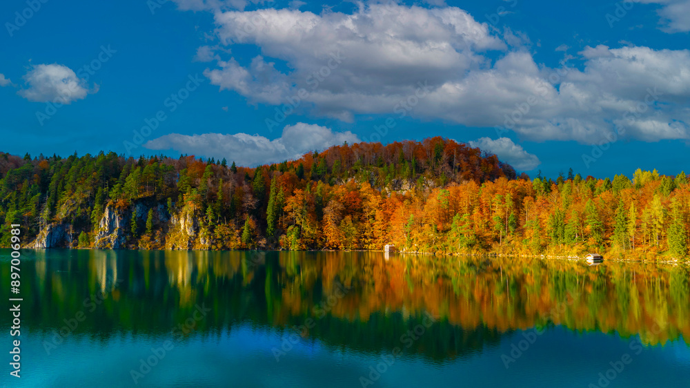 Panoramic view of the Alpsee lake in Autumn trees season and Bavaria Alps in background, beautiful reflections in water Panoramic view of the Alpsee lake in Autumn trees season and Bavaria Alps in background, beautiful reflections in water