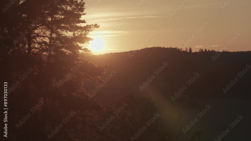 The sun sets over a hilly, forested landscape, casting a soft, golden light across the scene. The silhouette of the trees against the sky adds depth and tranquility to the image. The sun sets over a hilly, forested landscape, casting a soft, golden light across the scene. The silhouette of the trees against the sky adds depth and tranquility to the image.