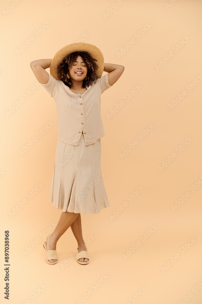 A stylish African American woman with curly hair is confidently posing in a summer dress and hat in a studio setting. A stylish African American woman with curly hair is confidently posing in a summer dress and hat in a studio setting.