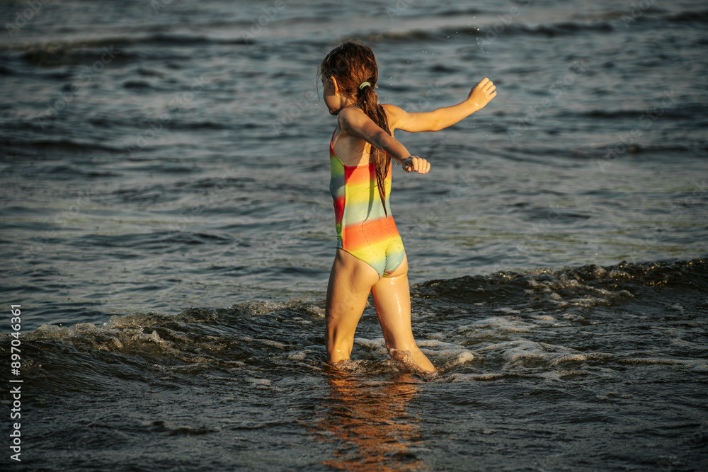 Naklejka premium A girl in a colorful swimsuit playing energetically in the ocean waves, capturing a moment of joy and movement by the sea. The image emphasizes the fun and excitement of a summer day at the beach