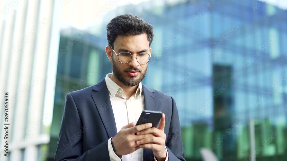 Confident businessman in suit stands near modern glass business office building holding smartphone in hands. Bearded man in glasses entrepreneur, executive or manager using a mobile phone outside