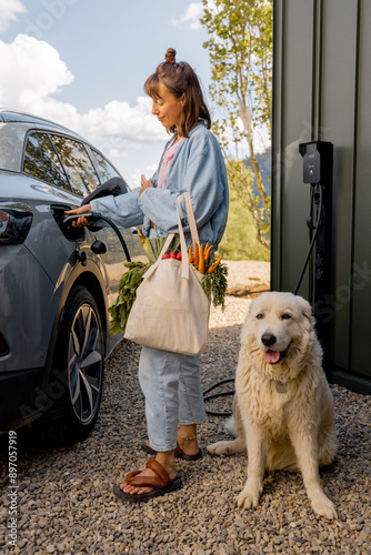 Young woman plugs a charger into electric vehicle standing with her cute white dog near her house outdoors. Concept of modern technologies, EV cars and friendship with pets