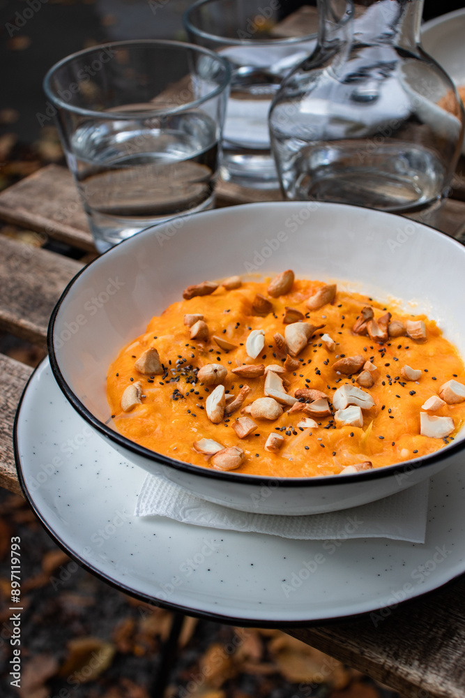 A traditional autumn dish of Ukrainian cuisine, boiled pumpkin porridge with nuts and chia seeds served with a glass of water on a wooden table outside, outdoors on the terrace. Close-up, vertical A traditional autumn dish of Ukrainian cuisine, boiled pumpkin porridge with nuts and chia seeds served with a glass of water on a wooden table outside, outdoors on the terrace. Close-up, vertical