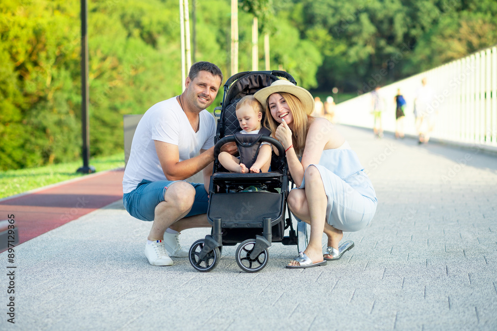 happy young parents mom and dad are walking with a baby in a stroller in the park in a hat in the summer in the setting sun and smiling having fun happy young parents mom and dad are walking with a baby in a stroller in the park in a hat in the summer in the setting sun and smiling having fun