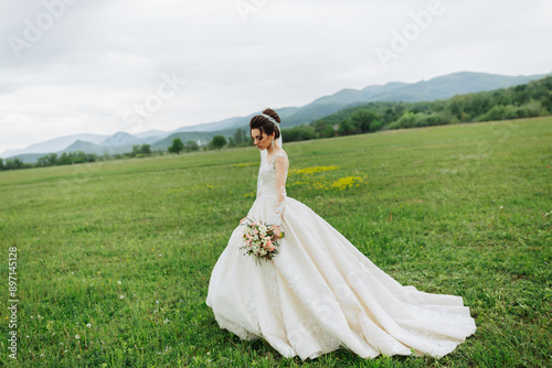 A bride is walking through a field with a bouquet of flowers. The scene is serene and peaceful, with the bride looking towards the camera. The grass is lush and green