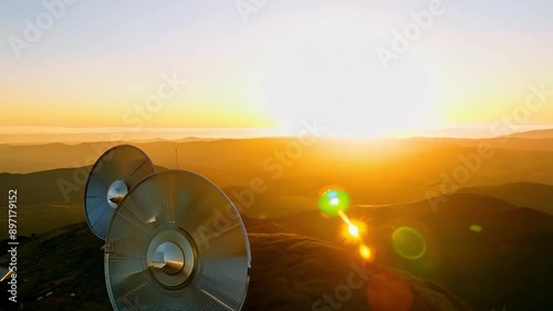 Two large, circular telescopes are mounted on a mountaintop, with the setting sun casting a golden glow over the landscape. photo