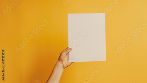 The image shows a hand holding a blank piece of paper against a yellow background.