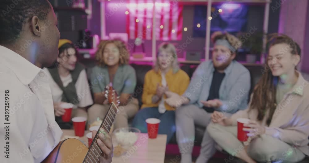 African American man playing the guitar while diverse group of friends singing laughing having fun in living room at night. Leisure activity and youth party concept. African American man playing the guitar while diverse group of friends singing laughing having fun in living room at night. Leisure activity and youth party concept.