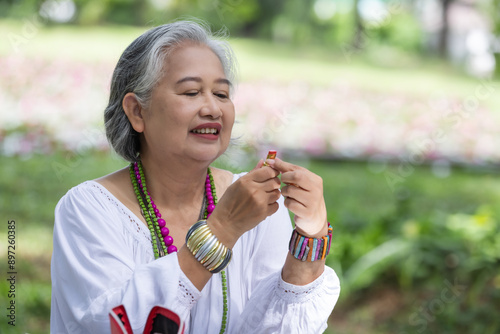 Elderly Woman Holding Lipstick and Smiling, Wearing Colorful Jewelry in a Garden Setting photo