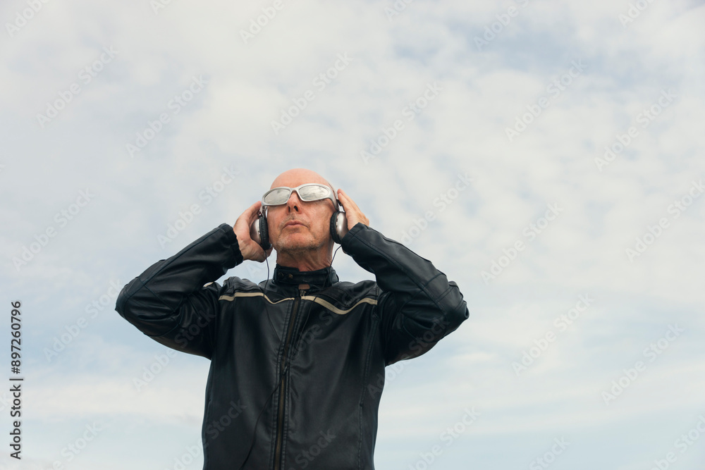 Attractive bald man with sunglasses and headphones. Outdoors with blue sky background Attractive bald man with sunglasses and headphones. Outdoors with blue sky background