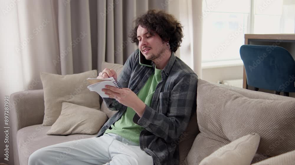 Confident brunette man with curly hair in a gray checkered shirt communicates on the phone and takes notes in a notepad at home on the sofa Confident brunette man with curly hair in a gray checkered shirt communicates on the phone and takes notes in a notepad at home on the sofa