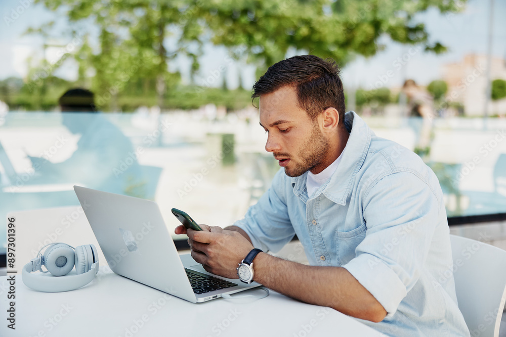 Young man focused on smartphone while working on a laptop in a modern outdoor café Young man focused on smartphone while working on a laptop in a modern outdoor café