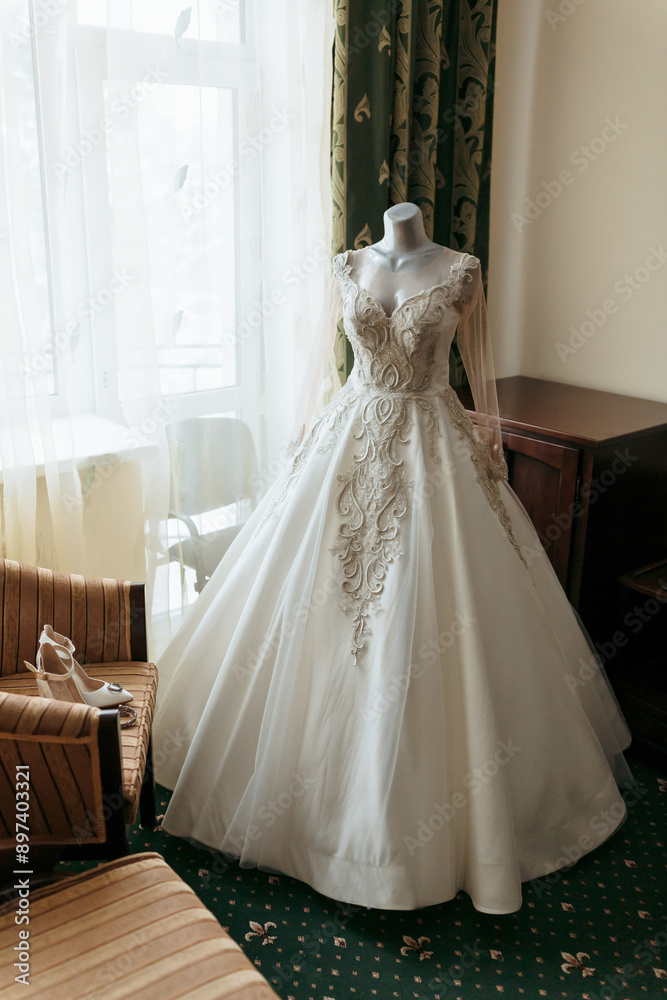 A white wedding dress is displayed on a mannequin in a room with a window. The dress is long and has a lace-like design