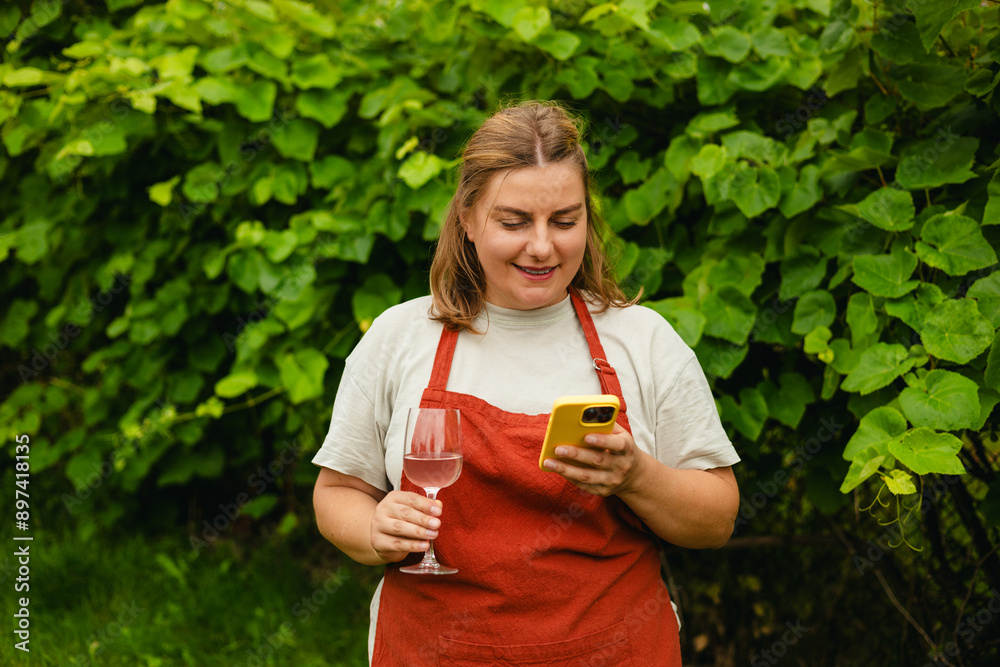 Woman with phone tasting white wine in vineyard. Portrait of pretty young woman holding glass of wine. Happy vintner drinks wine after successful grape harvesting, Natural wine industry. Harvest time Woman with phone tasting white wine in vineyard. Portrait of pretty young woman holding glass of wine. Happy vintner drinks wine after successful grape harvesting, Natural wine industry. Harvest time