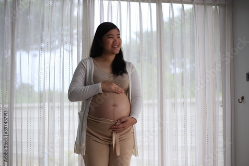 Happy pregnant woman with big tummy relax at home. Portrait of pregnant mature woman standing near the window and caresses her belly. Smiling woman expecting child and looking at camera. Free Download Happy pregnant woman with big tummy relax at home. Portrait of pregnant mature woman standing near the window and caresses her belly. Smiling woman expecting child and looking at camera. - GFXLoad