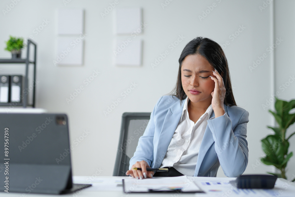 Tired asian businesswoman experiencing a headache, touching her head while sitting at her desk in a modern office Tired asian businesswoman experiencing a headache, touching her head while sitting at her desk in a modern office