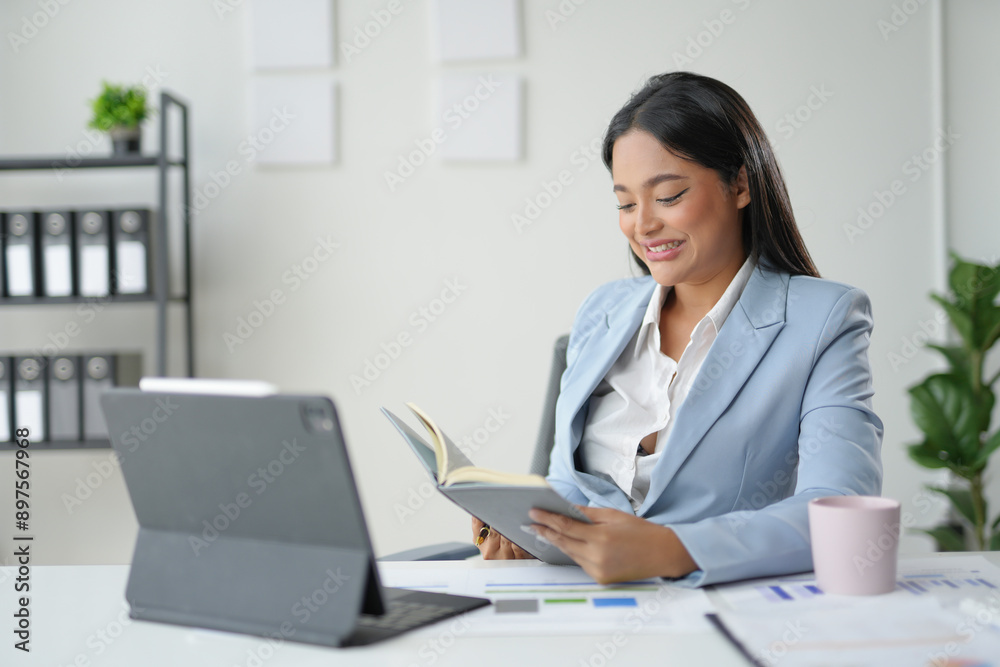 Businesswoman is sitting at her desk and comparing information from a book and a tablet Businesswoman is sitting at her desk and comparing information from a book and a tablet
