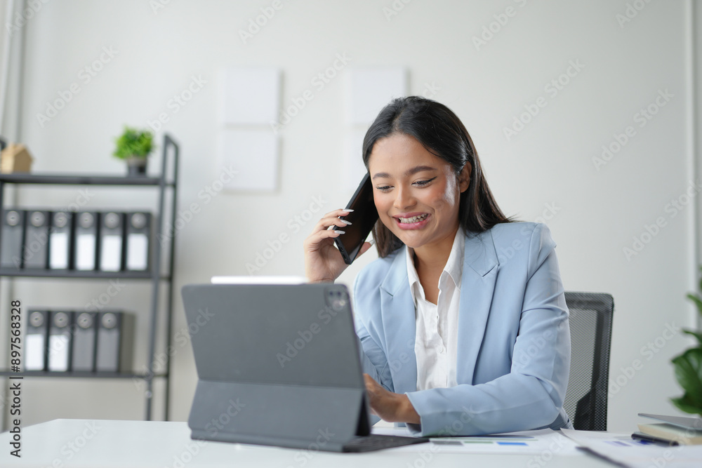 Young businesswoman is multitasking, smiling while talking on the phone and working on a tablet in her modern office