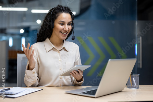 Smiling woman wearing headset participating in video call using tablet and laptop in modern office. Professional conducting virtual meeting with colleagues, engaging and communicating effectively.