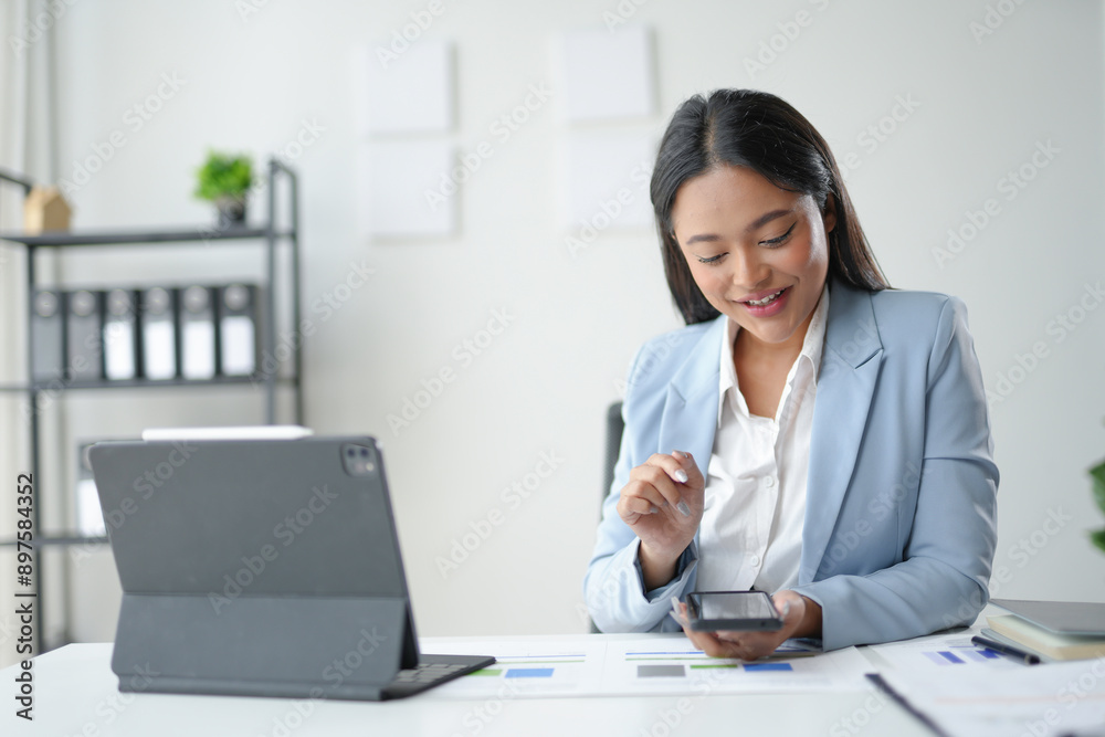 Smiling businesswoman analyzing financial charts on paperwork and using smartphone at office desk Smiling businesswoman analyzing financial charts on paperwork and using smartphone at office desk