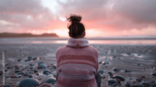 A touching image showcasing a person in a warm sweater sitting on a rocky beach, deep in reflection, with a stunning sunset sky creating a warm, tranquil ambiance. photo