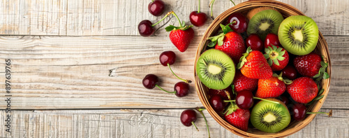A top-down view of a wooden basket filled with fresh organic fruits such as kiwis, strawberries, and cherries, with ample text space on a light wooden background.
