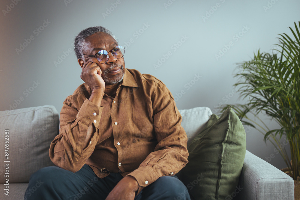 Fototapeta premium An elderly black man wearing glasses and a brown shirt looks pensive as he rests on a couch, his hand placed near his temple, indicating potential pain.