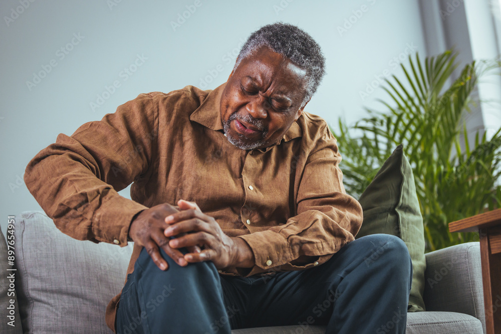 An older black man sits on a sofa, his expression tense with discomfort. He wears a casual shirt and appears to be in a living room setting. An older black man sits on a sofa, his expression tense with discomfort. He wears a casual shirt and appears to be in a living room setting.