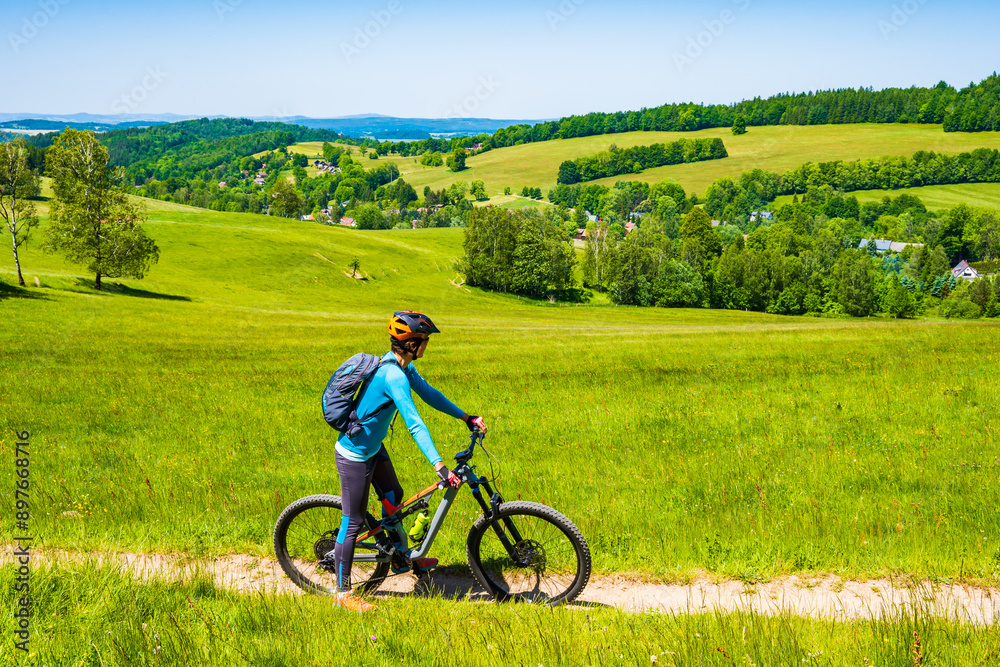 Fototapeta premium Woman with backpack riding mountain bike on rural road along green fields in Jizera Mountains, Singletrk pod Smrkem, Czech Republic