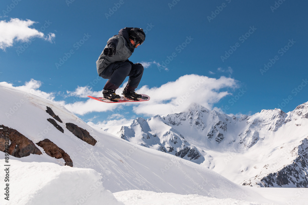Jumping snowboarder holds one hand on a snowboard against the background of a blue sky and large snow-capped mountains. Performing a stunt in flight Jumping snowboarder holds one hand on a snowboard against the background of a blue sky and large snow-capped mountains. Performing a stunt in flight