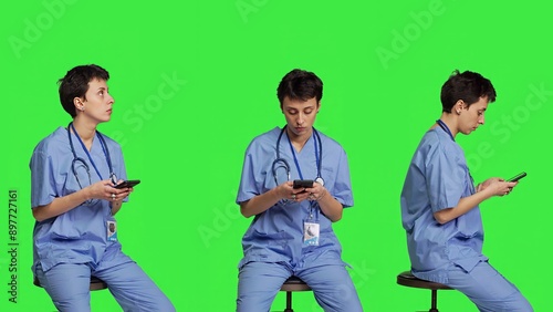 Nurse browsing online websites on smartphone app in studio, sitting on a chair against greenscreen backdrop. Medical specialist in scrubs using mobile phone and social media texting. Camera B.