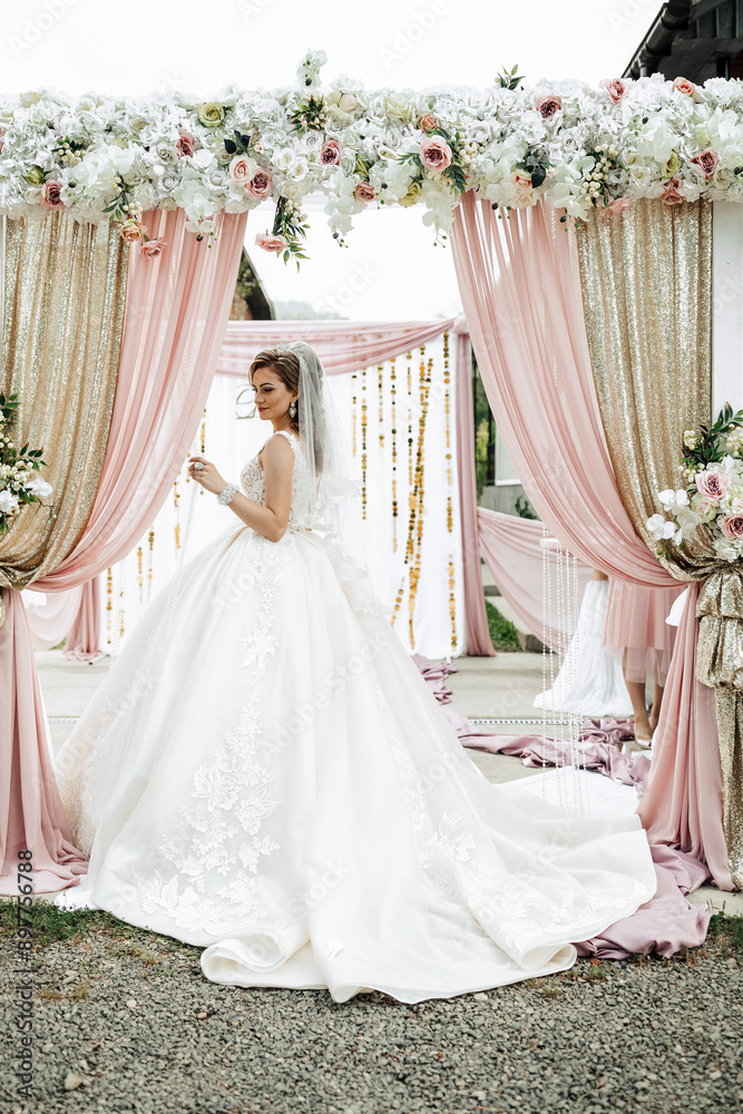 A woman in a white wedding dress stands in front of a pink archway. The archway is decorated with flowers and has a gold trim. The woman is wearing a veil and she is posing for a photo A woman in a white wedding dress stands in front of a pink archway. The archway is decorated with flowers and has a gold trim. The woman is wearing a veil and she is posing for a photo
