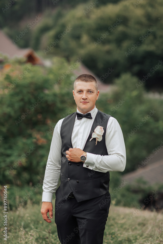 A man in a black vest and white shirt is standing in a field. He is wearing a black bow tie and holding a pink flower in his hand. Concept of elegance and sophistication