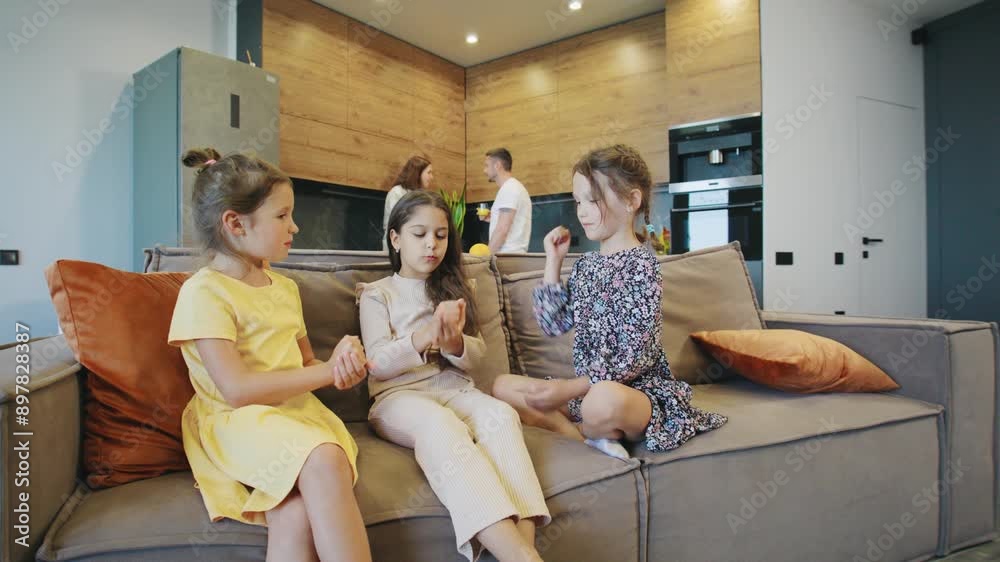 In a modern-style apartment, three girls are on the couch playing rock-paper-scissors. Their parents are visible in the background, engaged in household tasks, making for a lively and comfortable home In a modern-style apartment, three girls are on the couch playing rock-paper-scissors. Their parents are visible in the background, engaged in household tasks, making for a lively and comfortable home