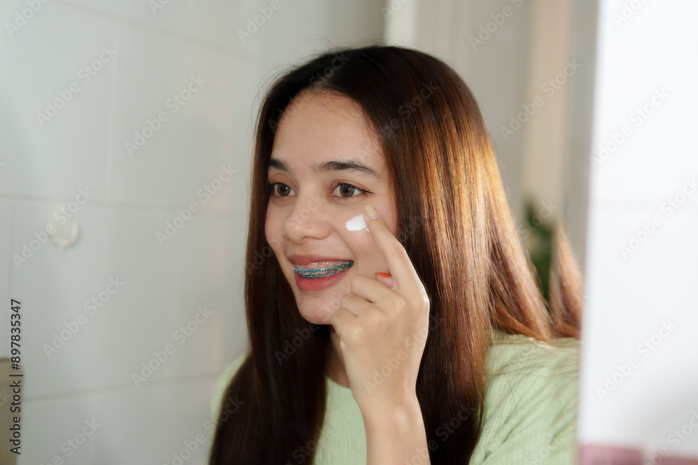 Asian teenage girl applying cream to cheek in bathroom. Long straight brown hair, light green ribbed top. Indoor setting with tiled walls. Expressing happiness and dedication to skincare routine. Asian teenage girl applying cream to cheek in bathroom. Long straight brown hair, light green ribbed top. Indoor setting with tiled walls. Expressing happiness and dedication to skincare routine.