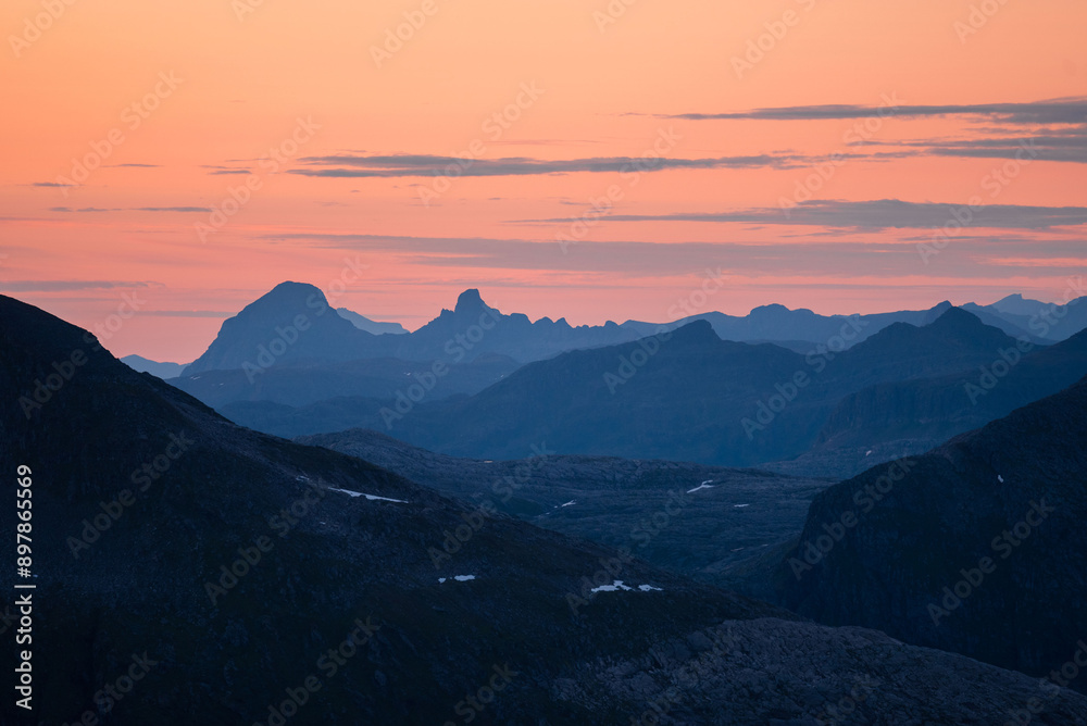 Midnight sun in arctic summer landscape of Norway. Silhouettes of mountain ranges in sunset colours of blue, orange and purple. Few clouds on clear dawn and dusk sky. Moody mountain range landscape Midnight sun in arctic summer landscape of Norway. Silhouettes of mountain ranges in sunset colours of blue, orange and purple. Few clouds on clear dawn and dusk sky. Moody mountain range landscape
