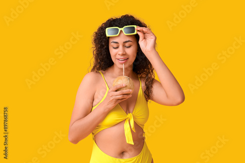 Beautiful young African-American woman with fresh coconut and straw on yellow background